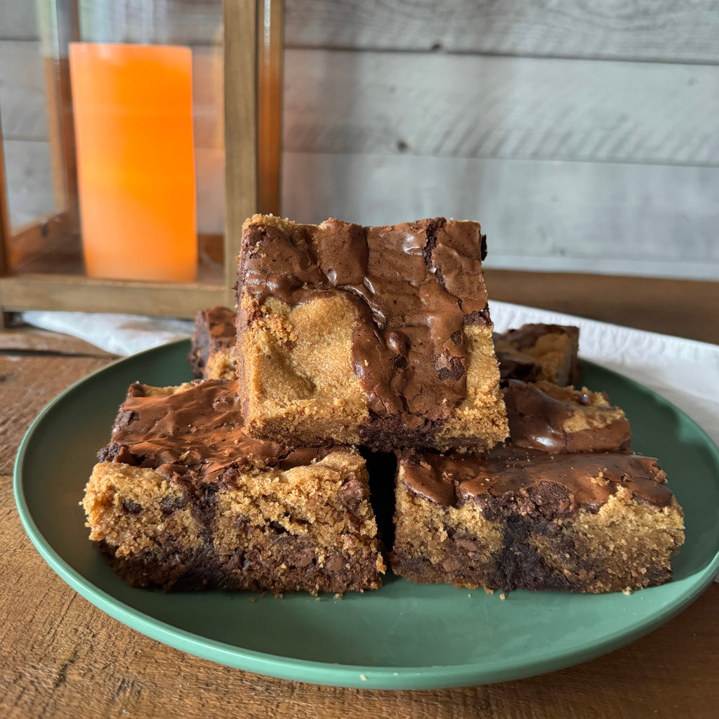 Four chocolate chip cookie brownies topped with chocolate drizzle, stacked on a green plate on a wooden table with a rustic lantern in the background.