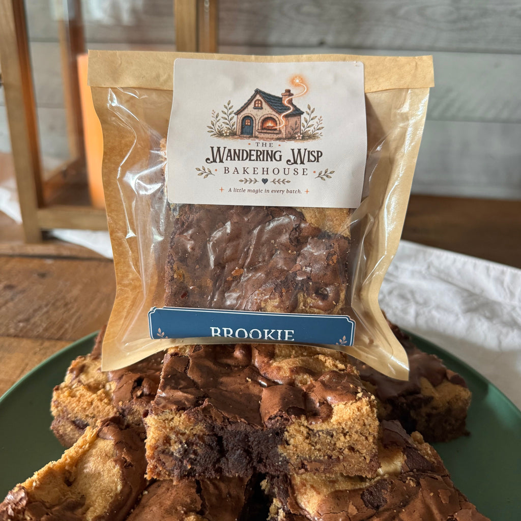 A bag of Wandering Wisp Bakehouse Brookie brownies sits on a green plate piled with cut pieces, with a rustic wooden table and paneled wall in the background.