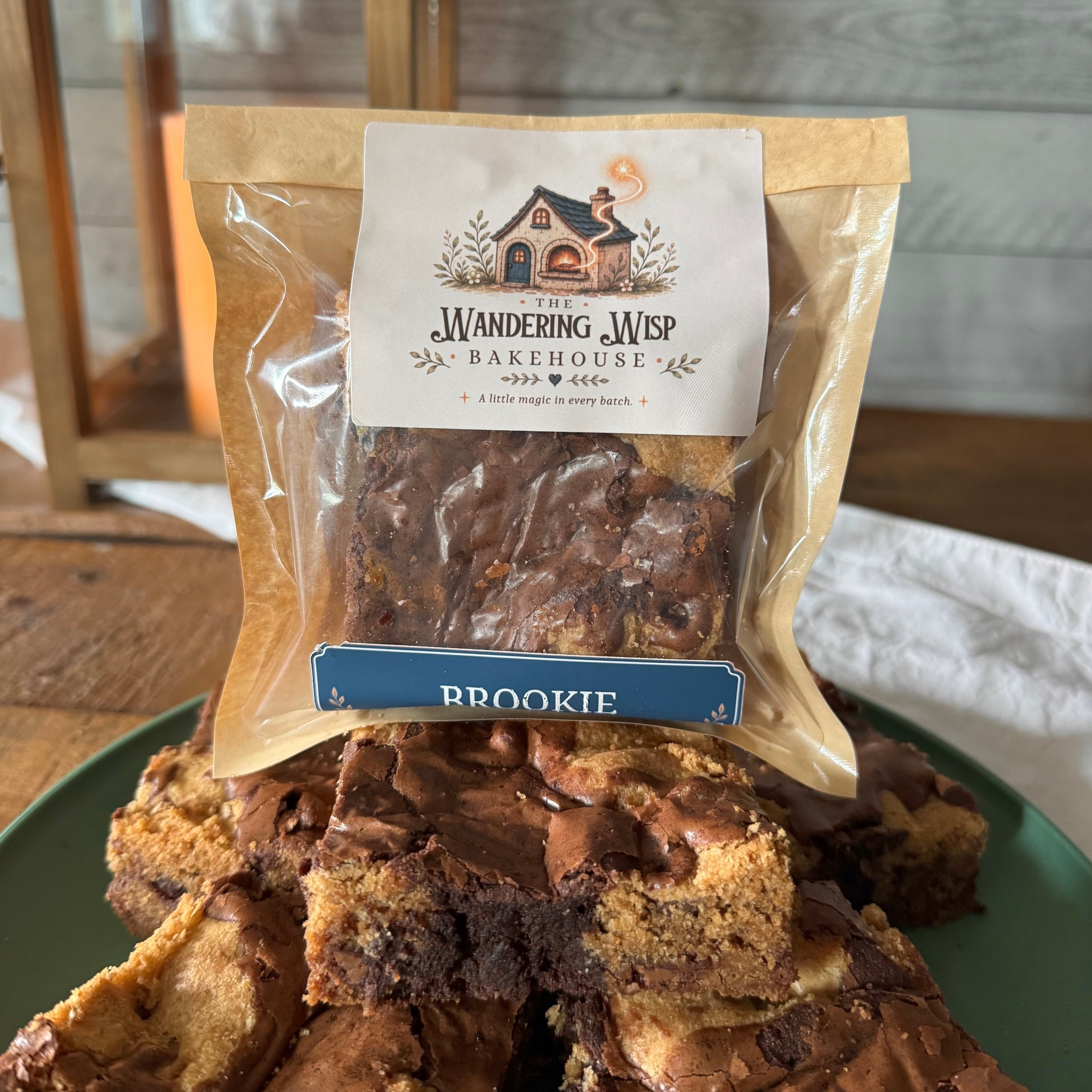 A bag of Wandering Wisp Bakehouse Brookie brownies sits on a green plate piled with cut pieces, with a rustic wooden table and paneled wall in the background.