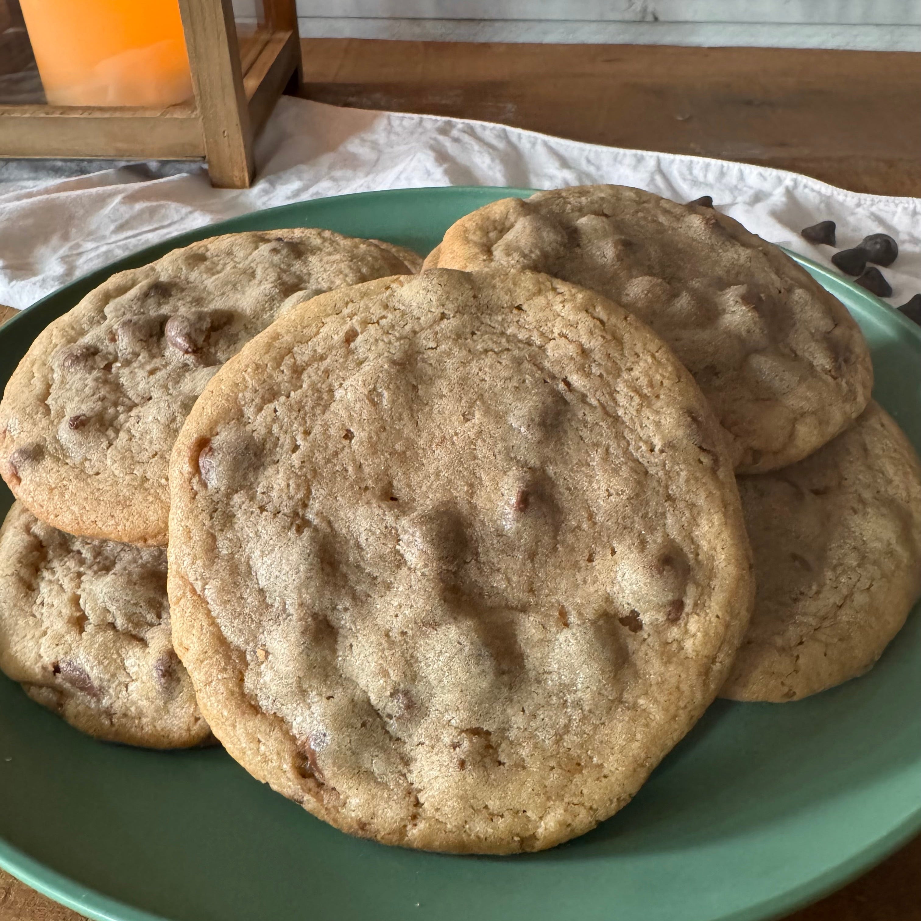 A plate of chocolate chip cookies on a green plate, set on a wooden table with a candle in the background.