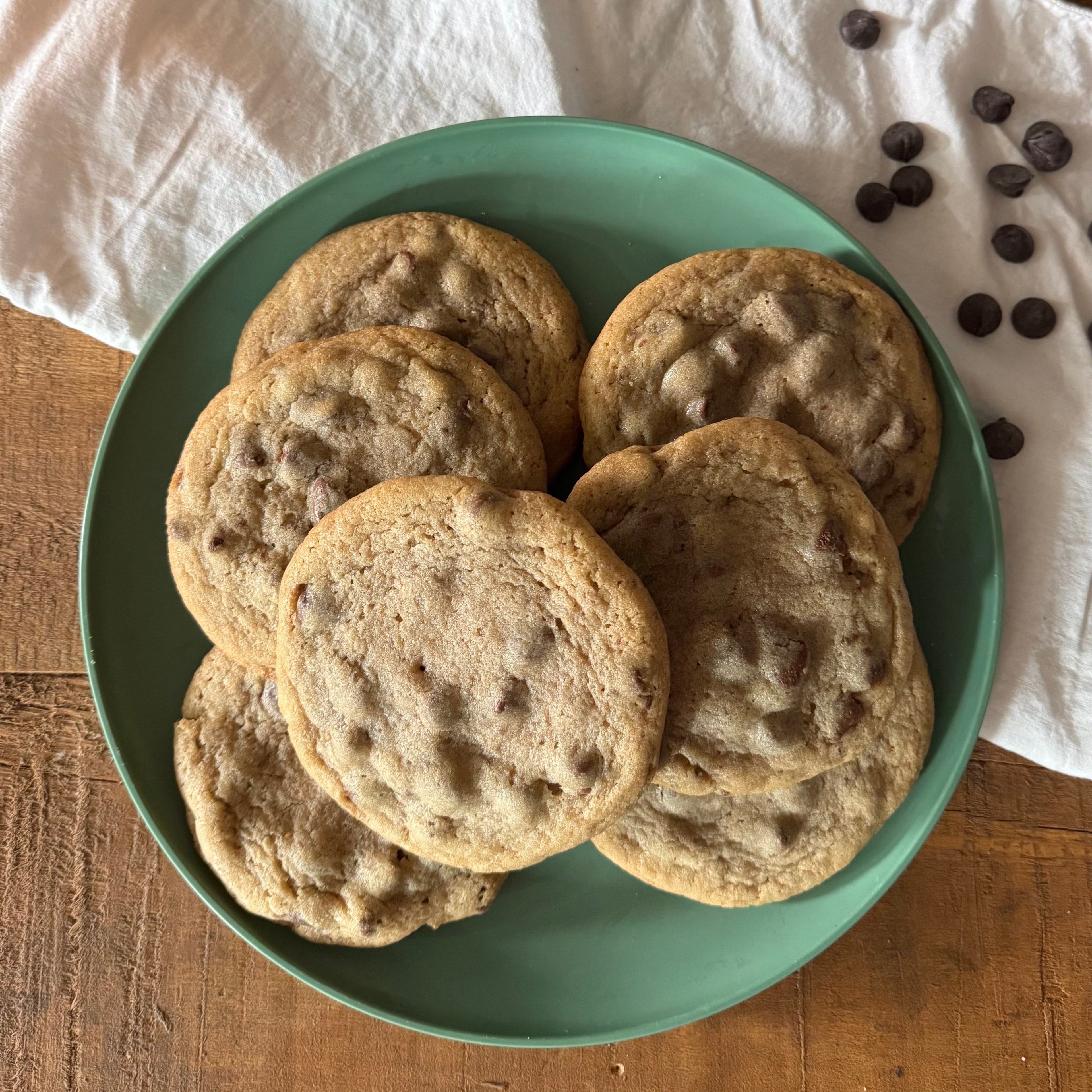 A green plate piled with chocolate chip cookies on a wooden table, with a white cloth and scattered chocolate chips in the background.