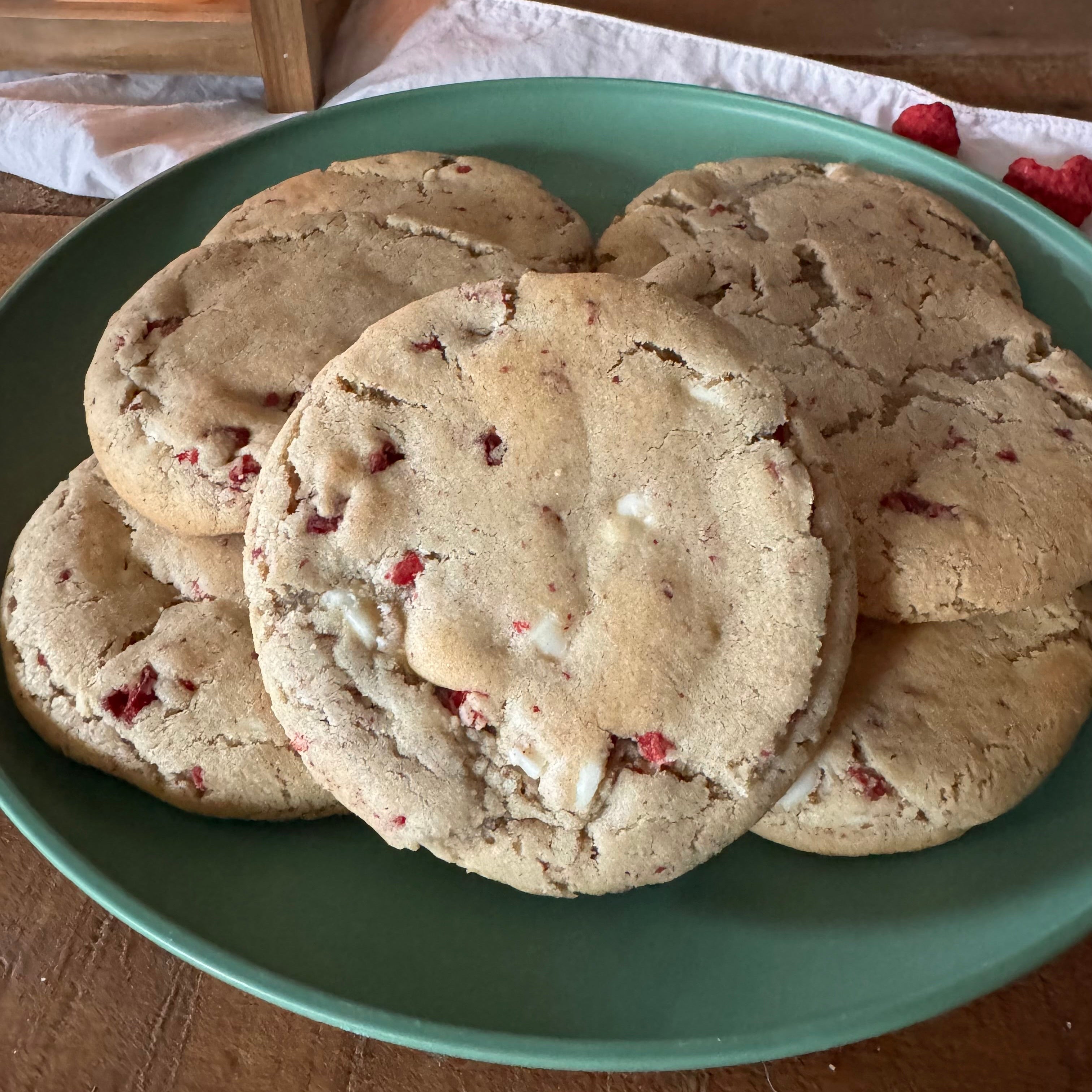 A stack of raspberry and white chocolate chip cookies on a green plate, set on a wooden table with a warm candle in the background.