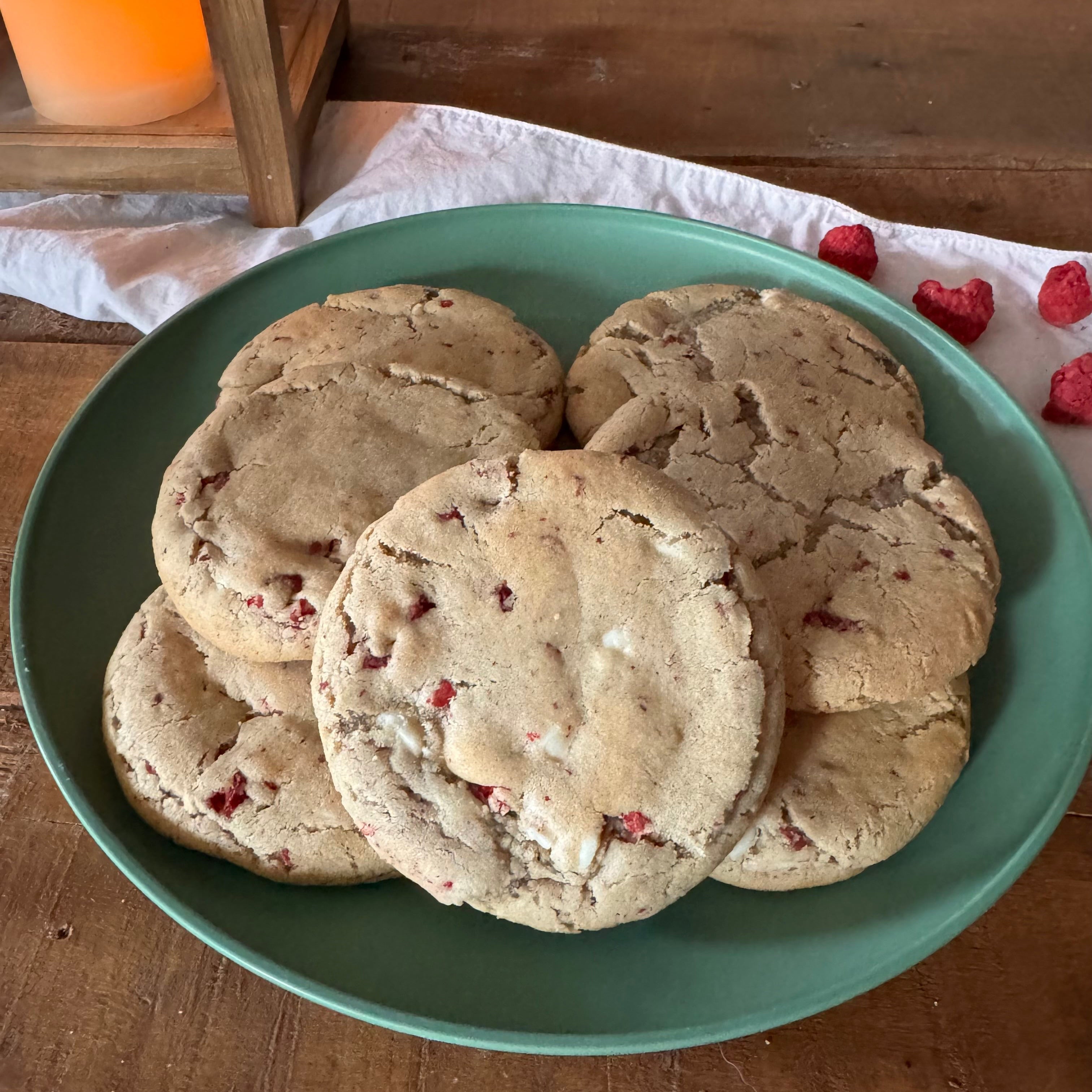 A green ceramic plate piled with raspberry cheesecake cookies set on a wooden table with a white cloth and raspberries nearby. 