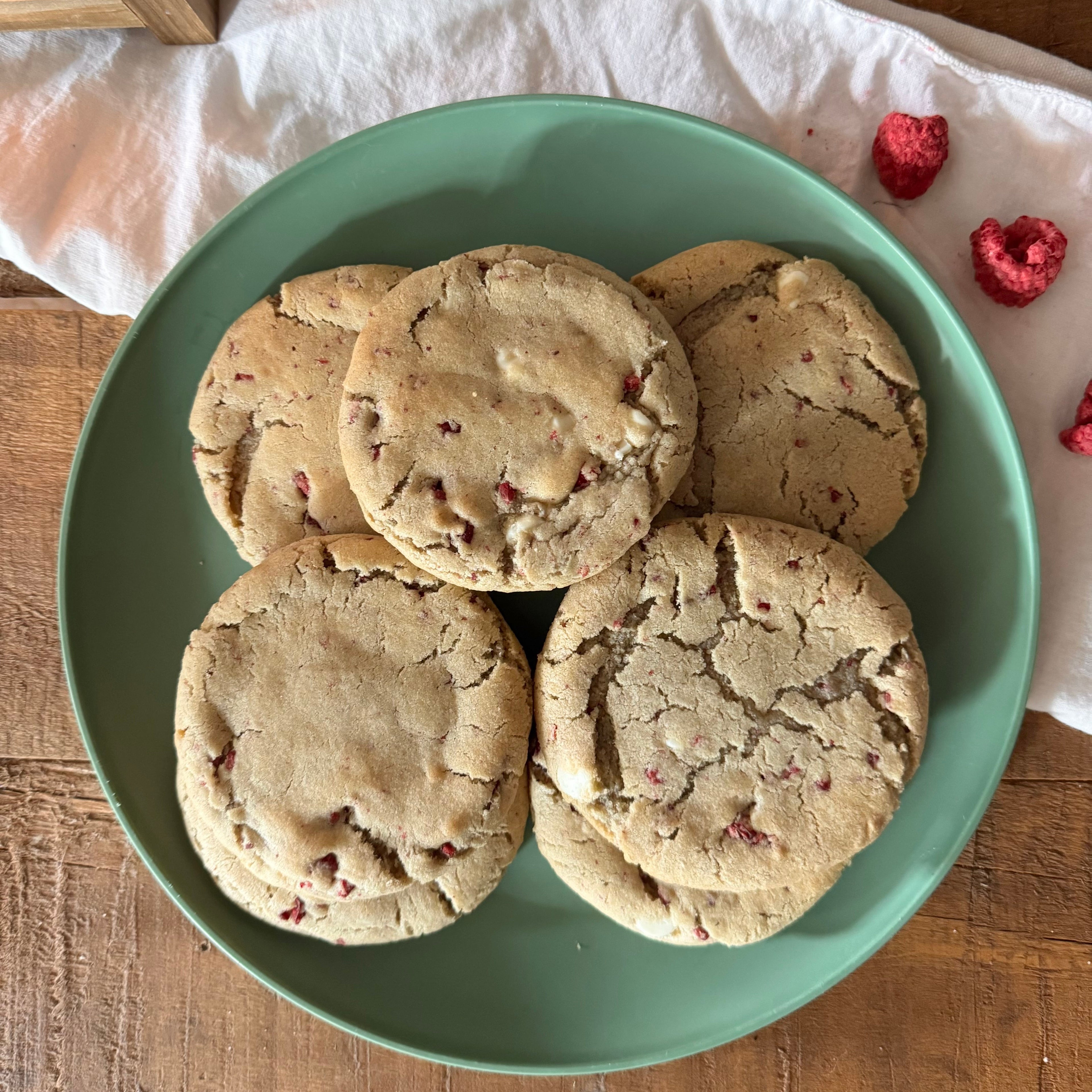 Raspberry cheesecake cookies stacked on a mint green plate over a wooden table with a white cloth and raspberries nearby.