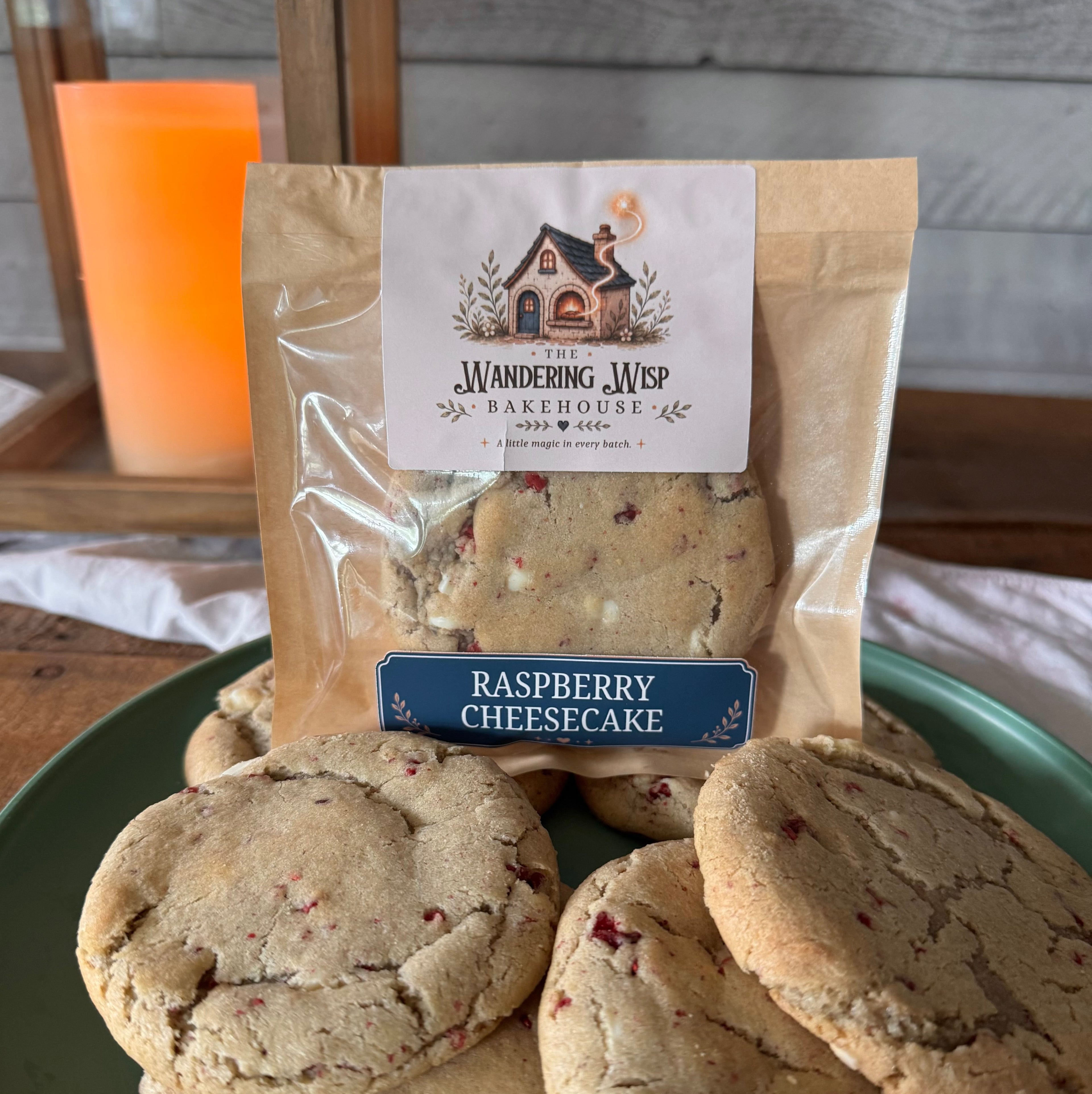 A bag of Wandering Web Bakery Raspberry Cheesecake cookies displayed on a green plate with several cookies stacked in front, set against a rustic wooden background with a glowing orange candle.