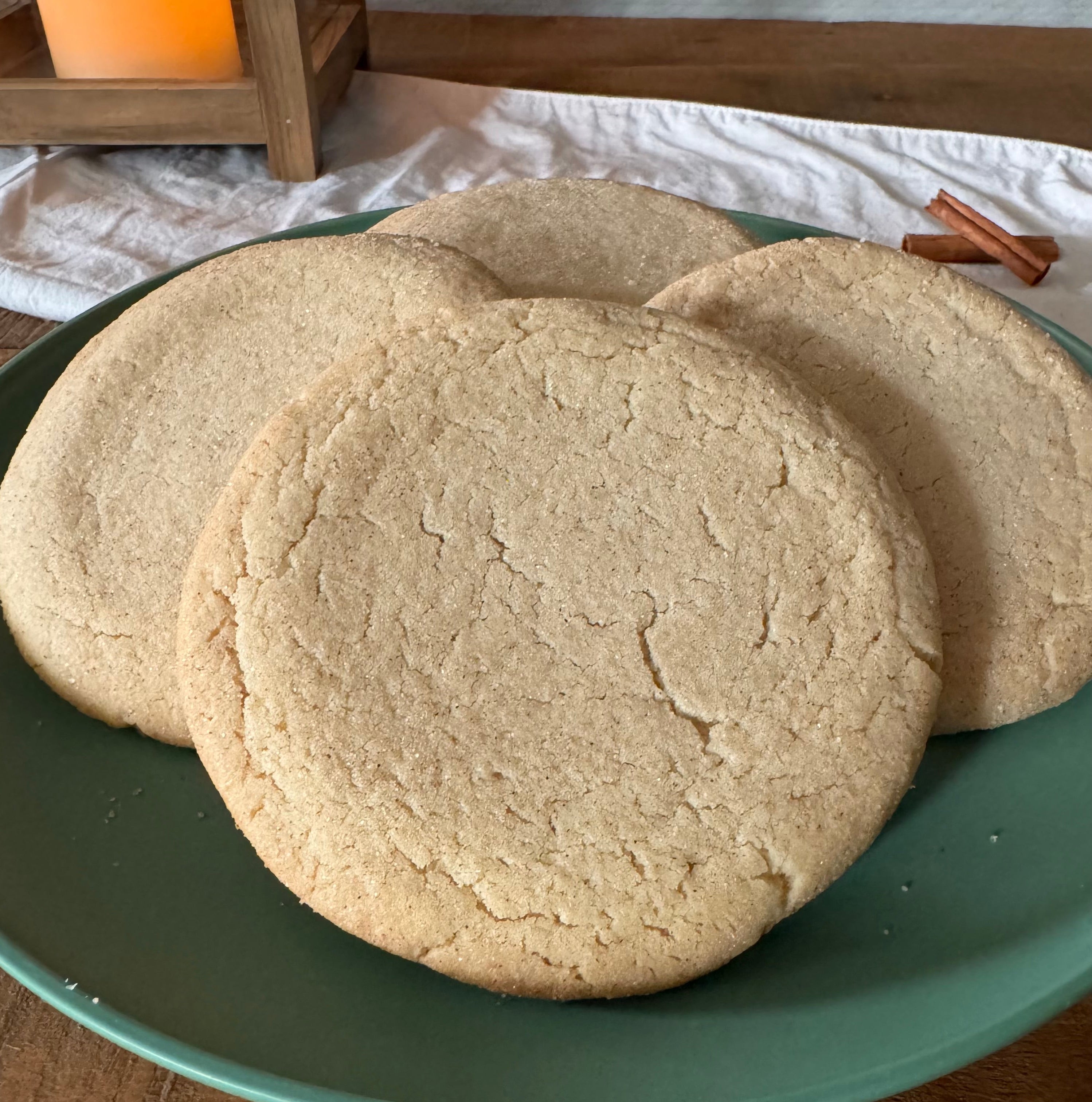 Three round snickerdoodle cookies on a green plate, set on a wooden table with a white cloth and a warm candle in the background.