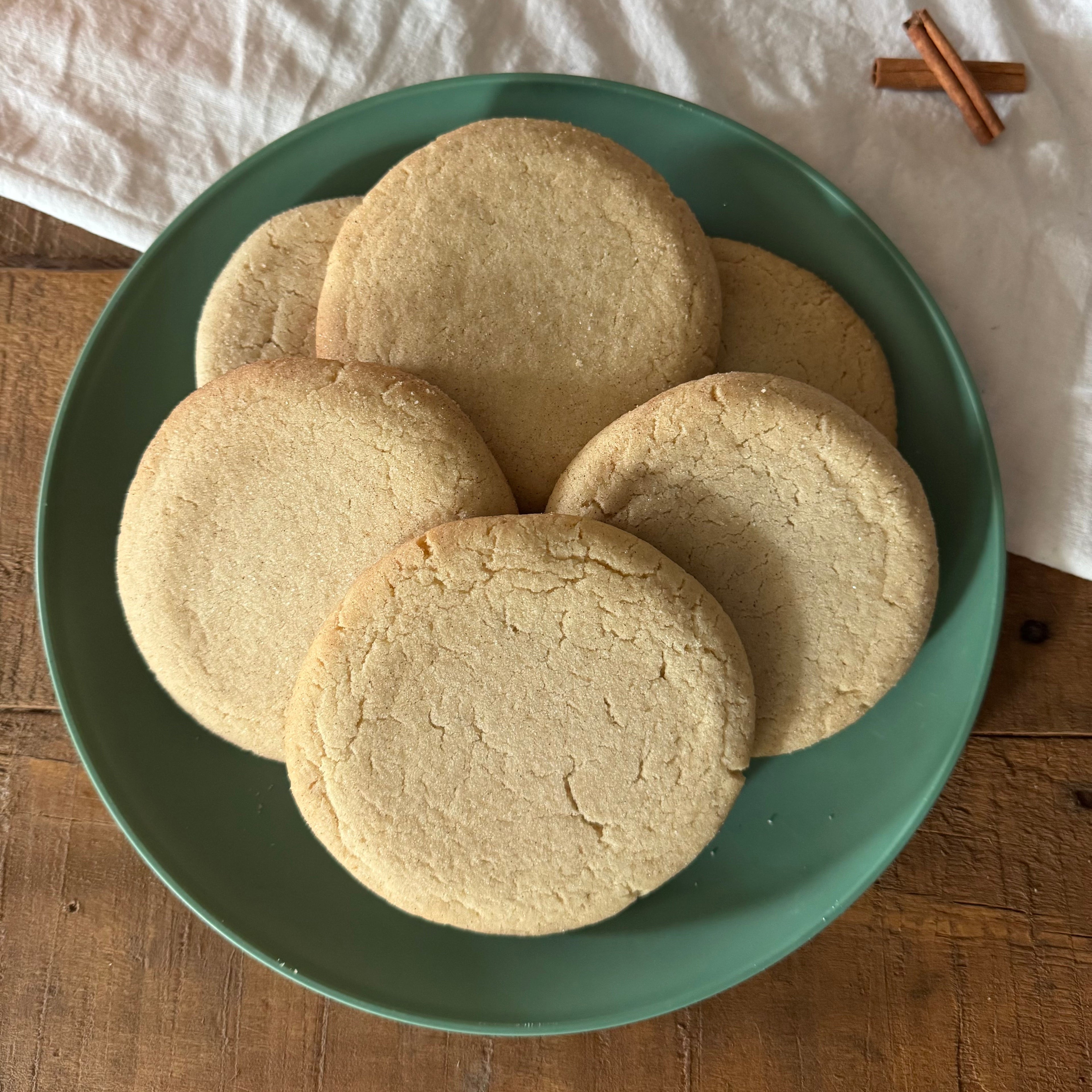 Six snickerdoodle cookies stacked on a green ceramic plate, set on a rustic wooden table next to a white cloth and cinnamon sticks.