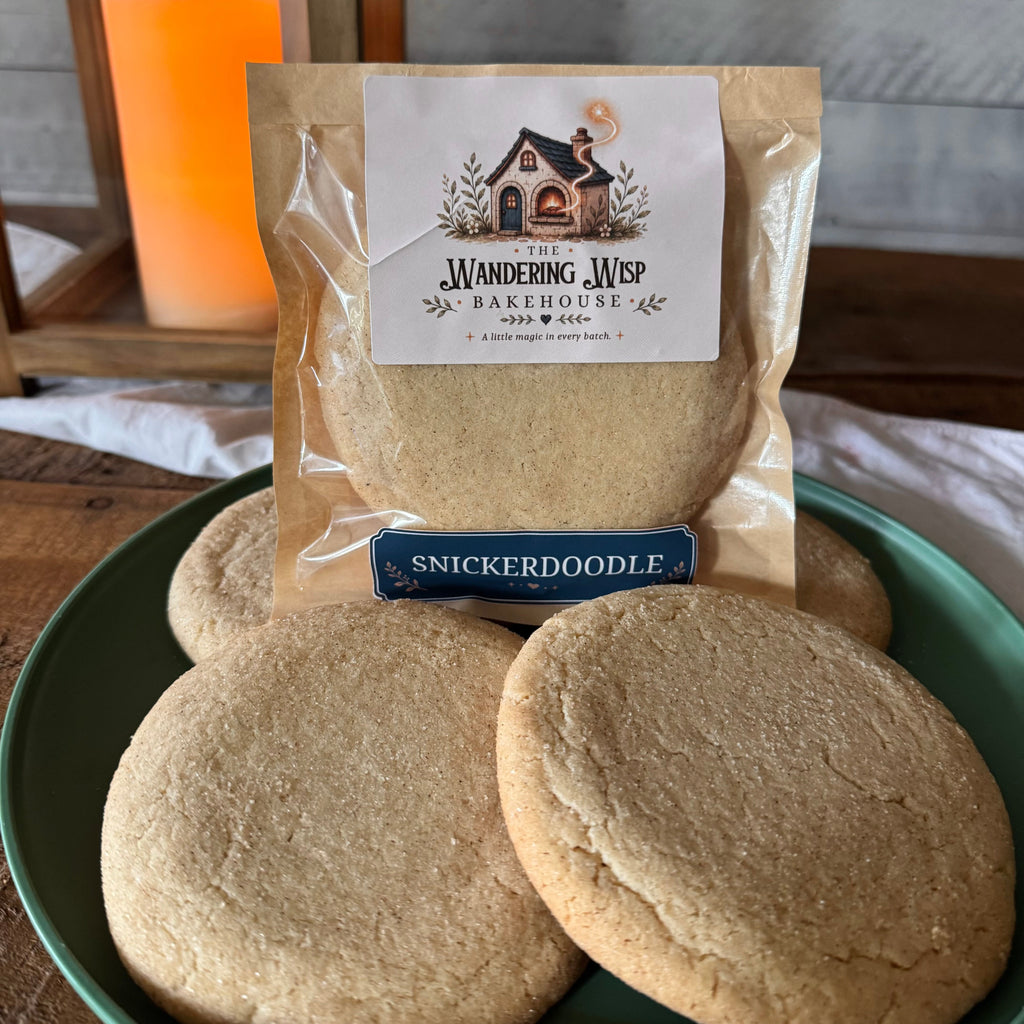 A package of Snickerdoodle cookies from The Wandering Web Bakehouse sits on a green plate with several baked cookies in front, set on a wooden table with a blurred candle in the background.