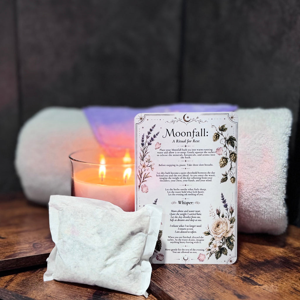 Bath products including a candle, ritual card and bath tea bag on a wooden shelf by a bathtub.